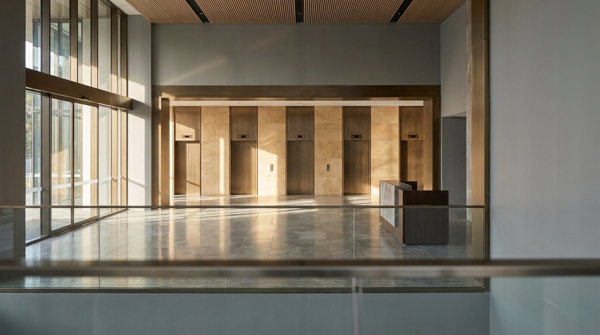 A double-height commercial office building lobby with a bank of lift doors and an arrivals floor seen from the mezzanine